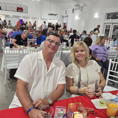 Two guests seated at Taste of Milam with event tables and community supporters in the background.